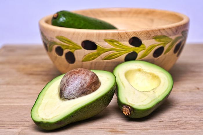 Green sliced avocado on a wooden tabletop.  An unsliced avocado in a wooden bowl with a green and purple leaf design sits behind it.