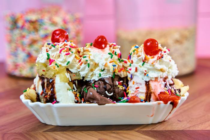 A banana sunday with three scoops of ice cream in a long white bowl.  There's vanilla, chocolate, and strawberry ice cream with whipped cream and cherries on top.  It is shown in front of a bright pink background.